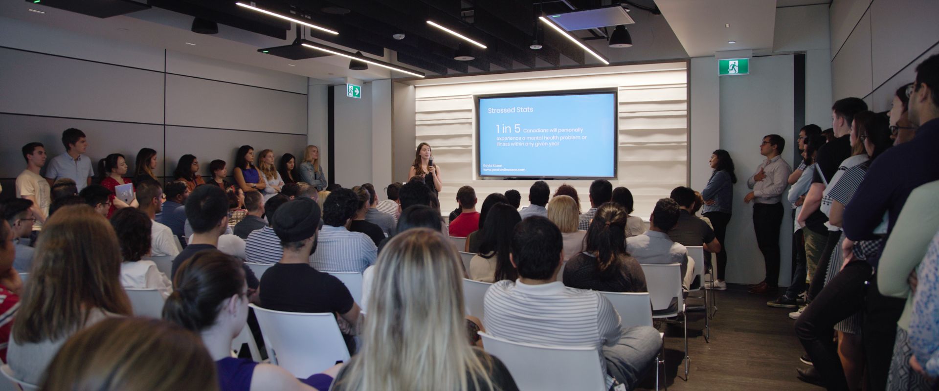 Room full of people standing against wall and sitting in chairs as they listen to Twello's CEO, Kayla Baum, do a presentation.