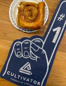 A cinnamon bun sits on a paper plate wth a Cultivator foam finger laying next to it.