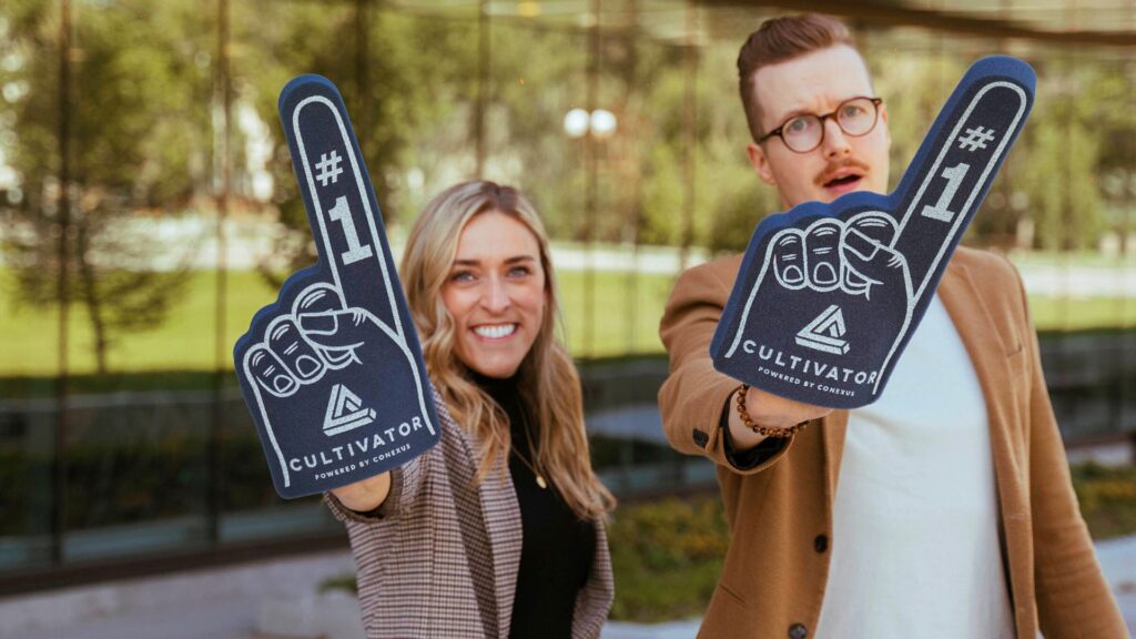 A woman and man raise their hands towards the camera with a Cultivator foam finger pointing upwards cheering.