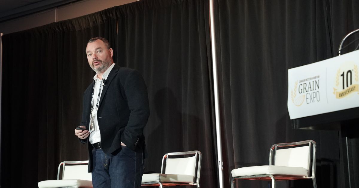 Jason McNamee stands on the Grain Expo stage with white tall stools behind him and the Grain Expo podium positioned at the side of the stage.