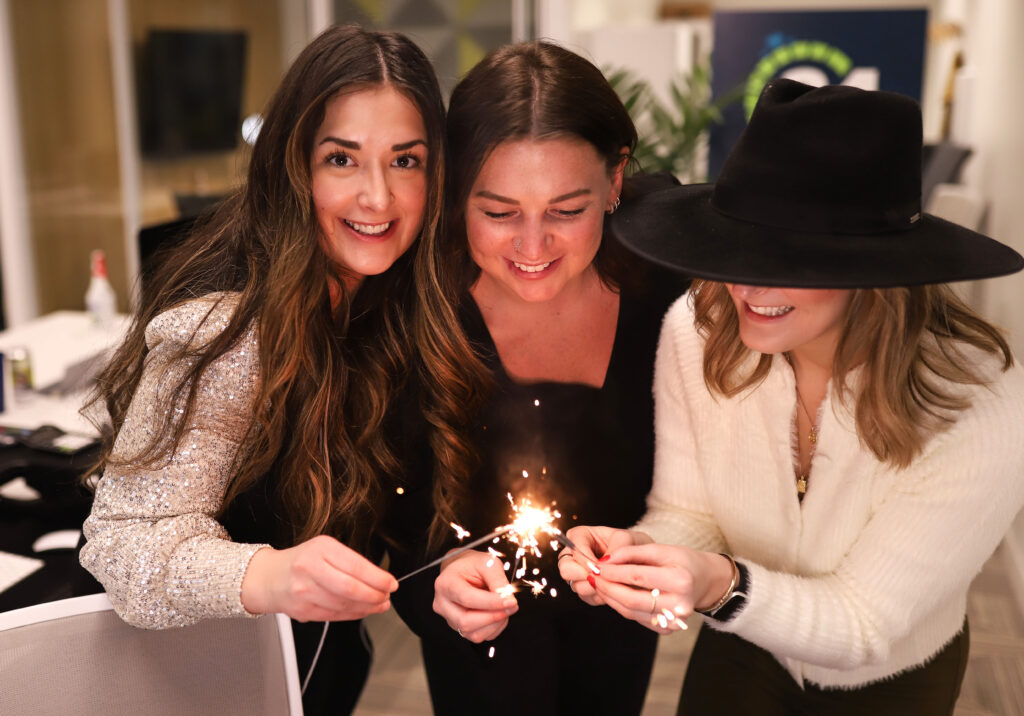 Ami Caragata, Bre Walkeden, and Breanne Dmytriw stand close together lighting and holding birthday candle sparklers.