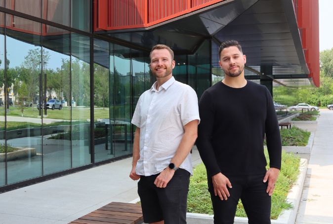 Two males standing side by side in front of a glass building