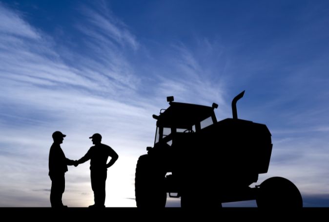 TWO FARMERS STAND NEAR A TRACTOR SHAKING HANDS.