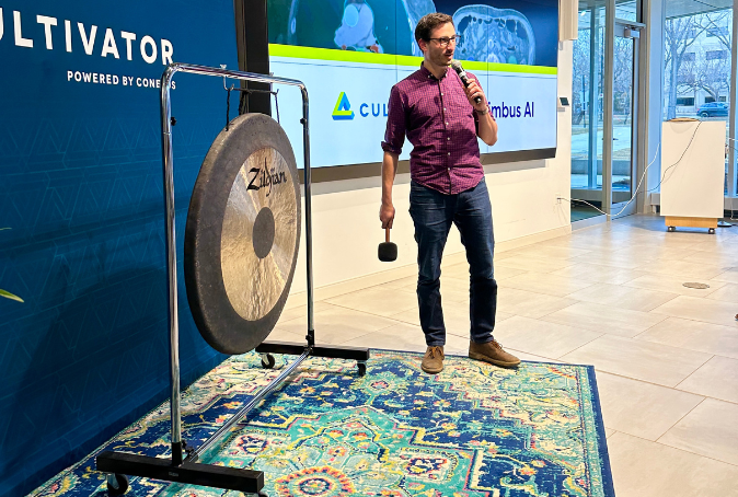 Man in a red shirt stand next to a large gong while holding a soft padded mallet and a microphone.