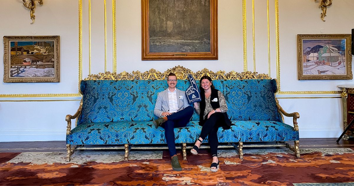 Bre Walkeden and Jordan McFarlen sit on a fancy teal blue damask patterned couch with gold detail. Bre Walkeden is holding a Cultivator foam finger in the air.