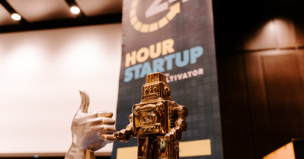 A thumbs up statue and gold robot statue in front of 24 Hour Startup banner