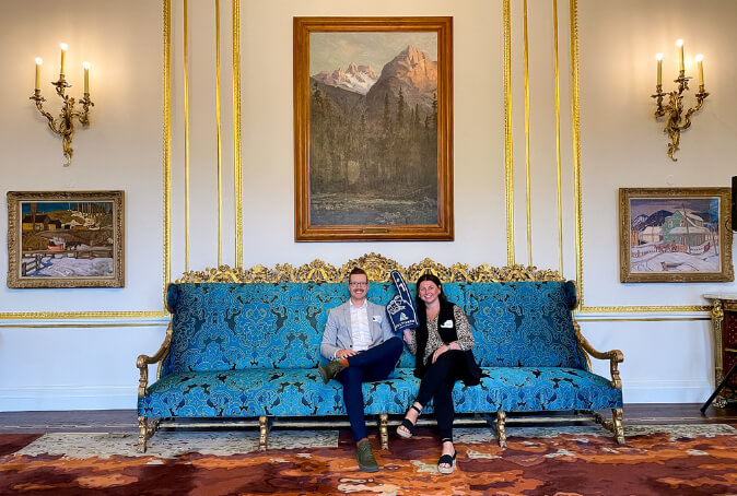 Bre Walkeden and Jordan McFarlen sit on a fancy teal blue damask patterned couch with gold detail. Bre Walkeden is holding a Cultivator foam finger in the air.
